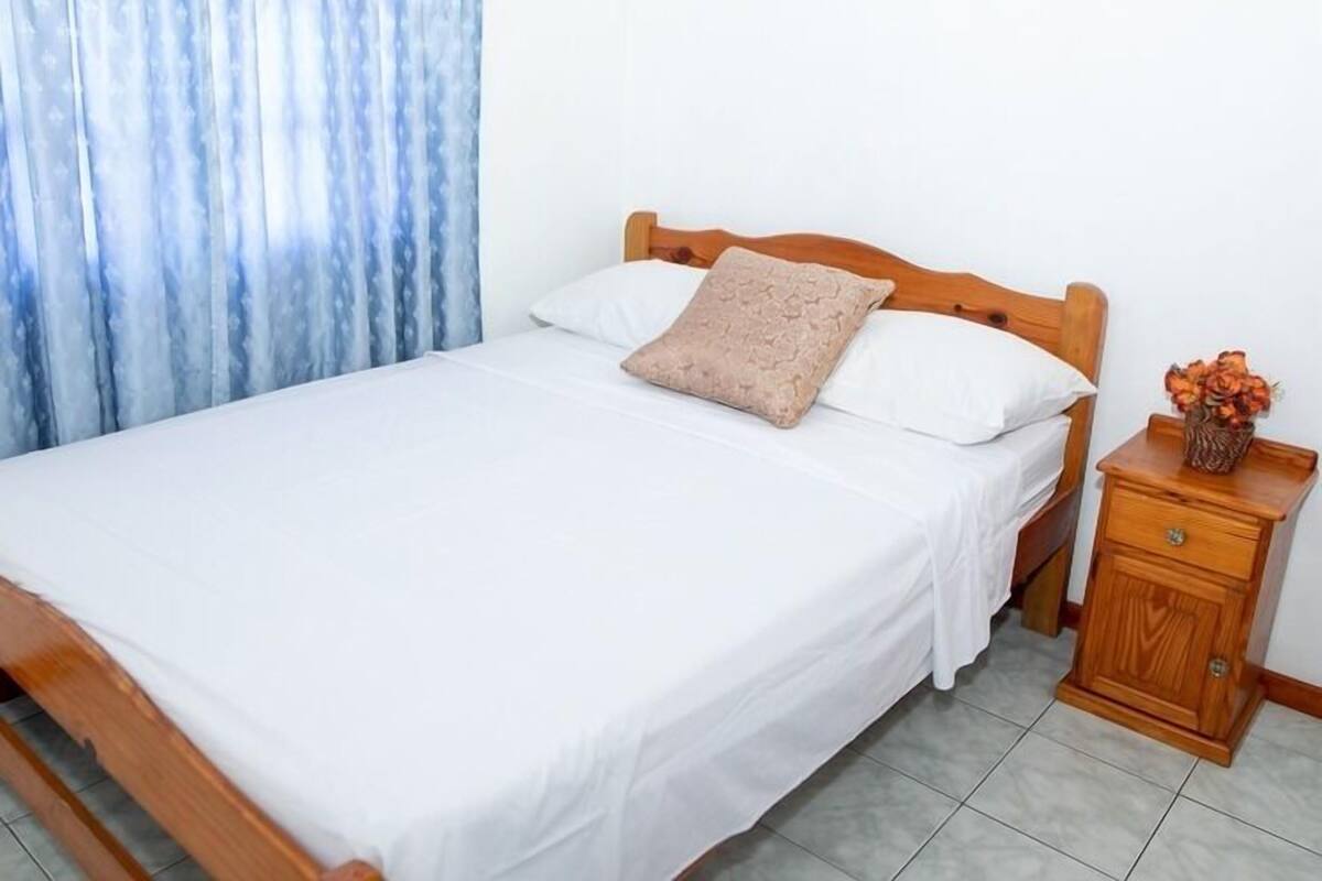 A neatly made bed with white bedding and a wooden headboard, a wooden nightstand with a vase of flowers, and a blue curtain partially drawn to the side.