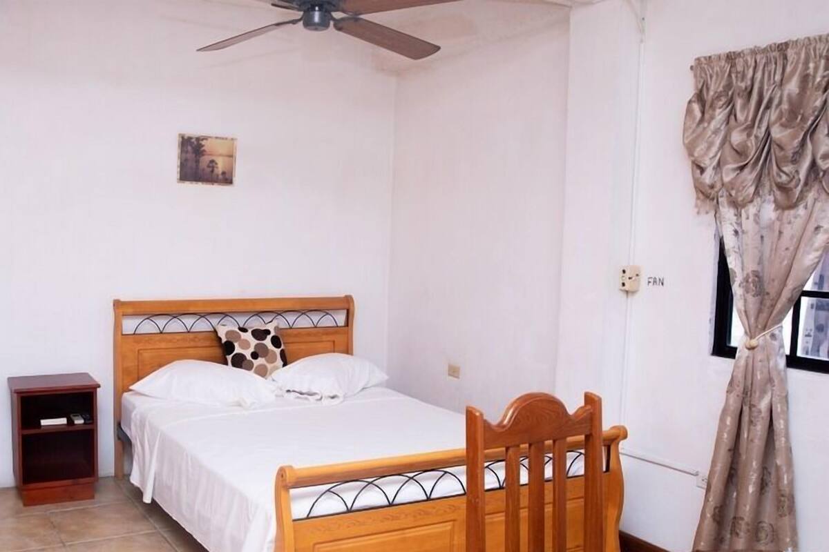 A bedroom with a wooden bed, a nightstand, a chair, a ceiling fan, and a window with curtains.