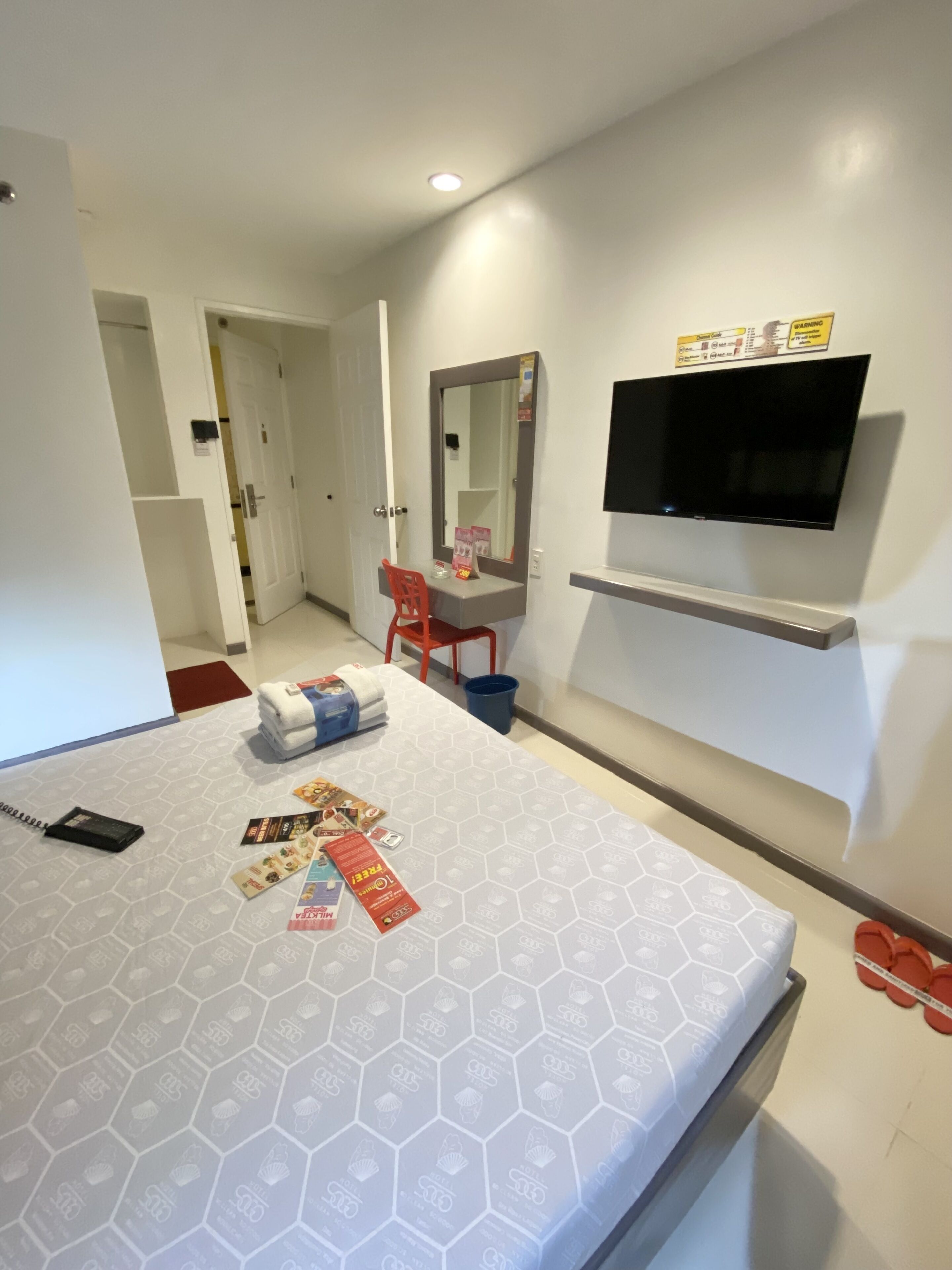 A hotel room with a bed, a TV mounted on the wall, a small table with toiletries, and a mirror above the table.