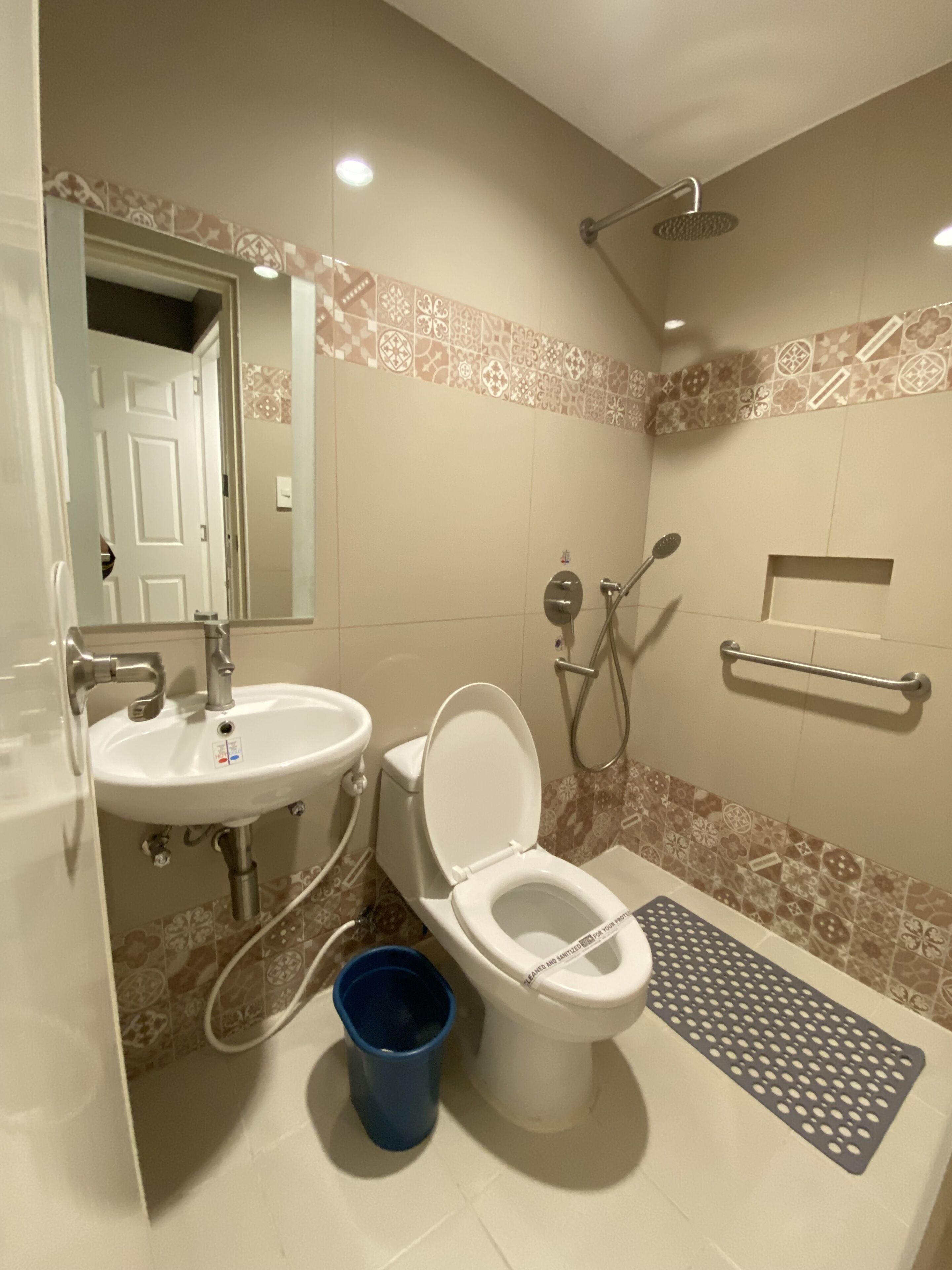 A bathroom with a white sink, toilet, and shower. There is a blue trash bin and a towel rack on the wall.