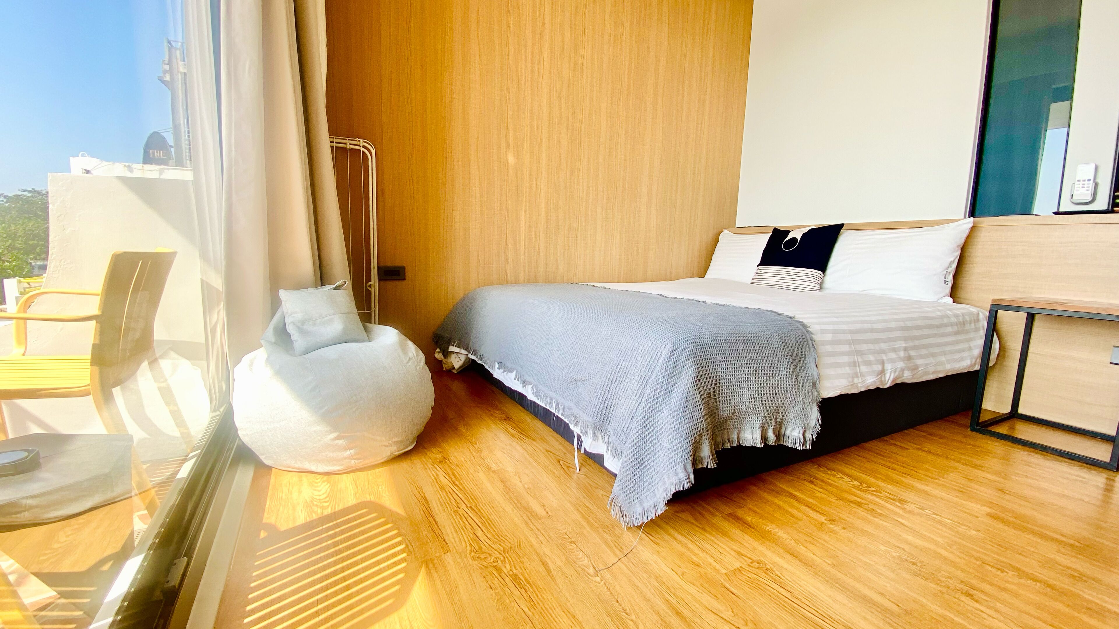 Een moderne slaapkamer met een houten vloer, een bed met een grijze deken, een witte pluinfauteuil en een houten nachtkastje.