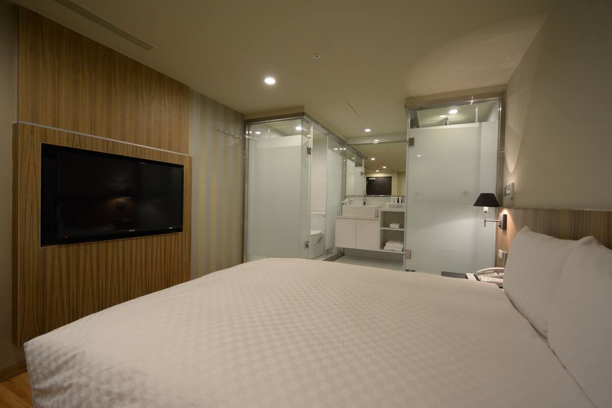 A hotel room with a large bed, a flat-screen TV mounted on the wall, a bathroom with a glass shower enclosure, and a small seating area with a table and chairs.