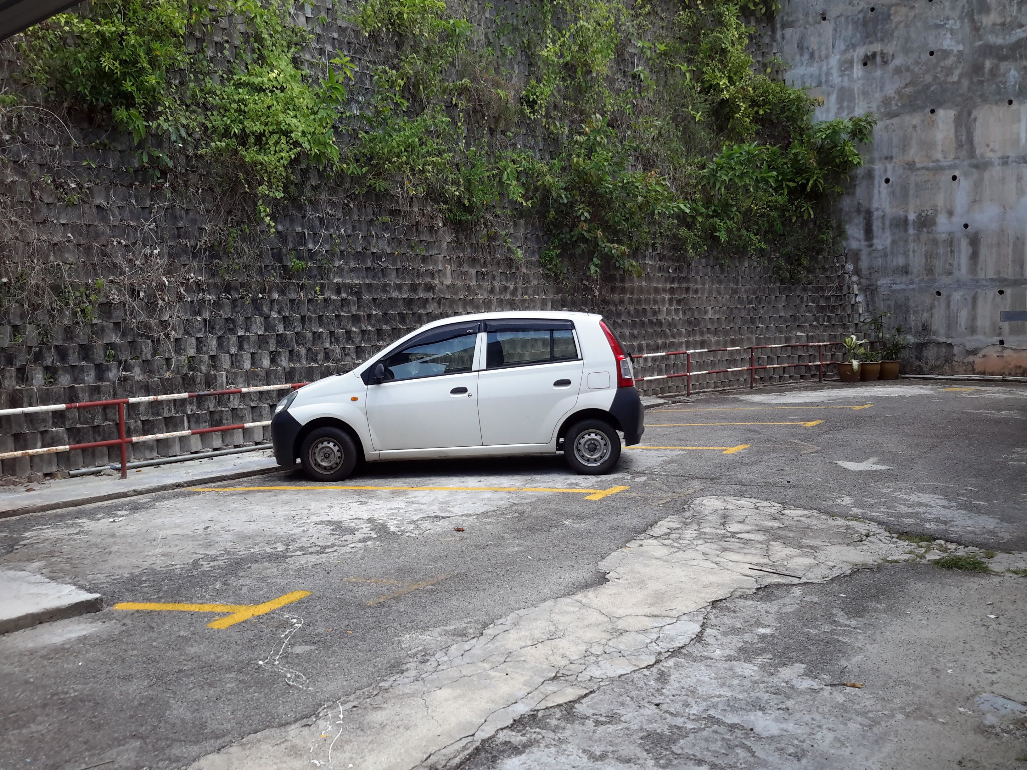 Une voiture blanche est garée sur une route défoncée, avec un mur de briques et de la verdure en arrière-plan.