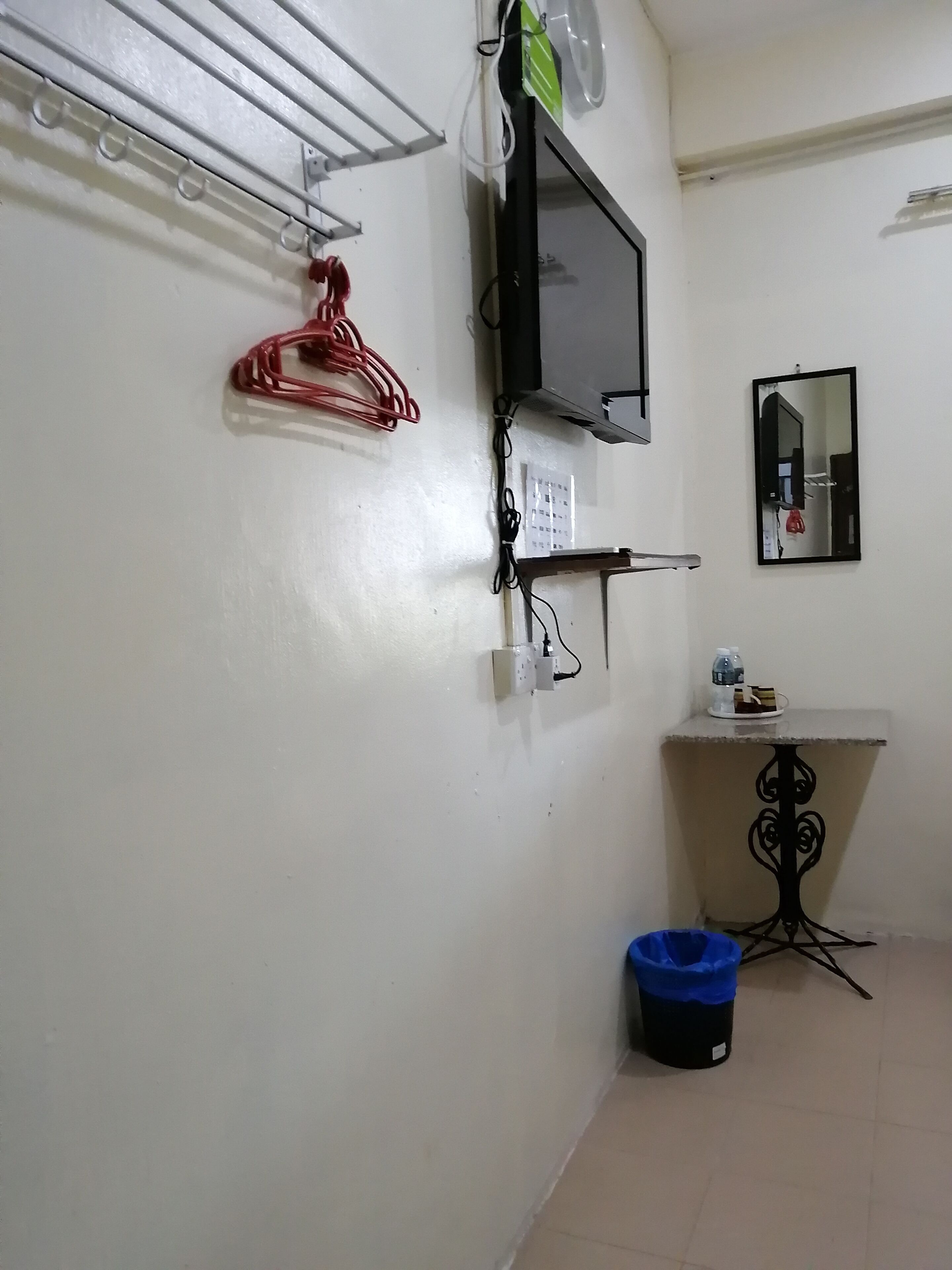 A small room with a TV mounted on the wall, a mirror, a shelf with a bottle and a plate, and a blue trash bin.
