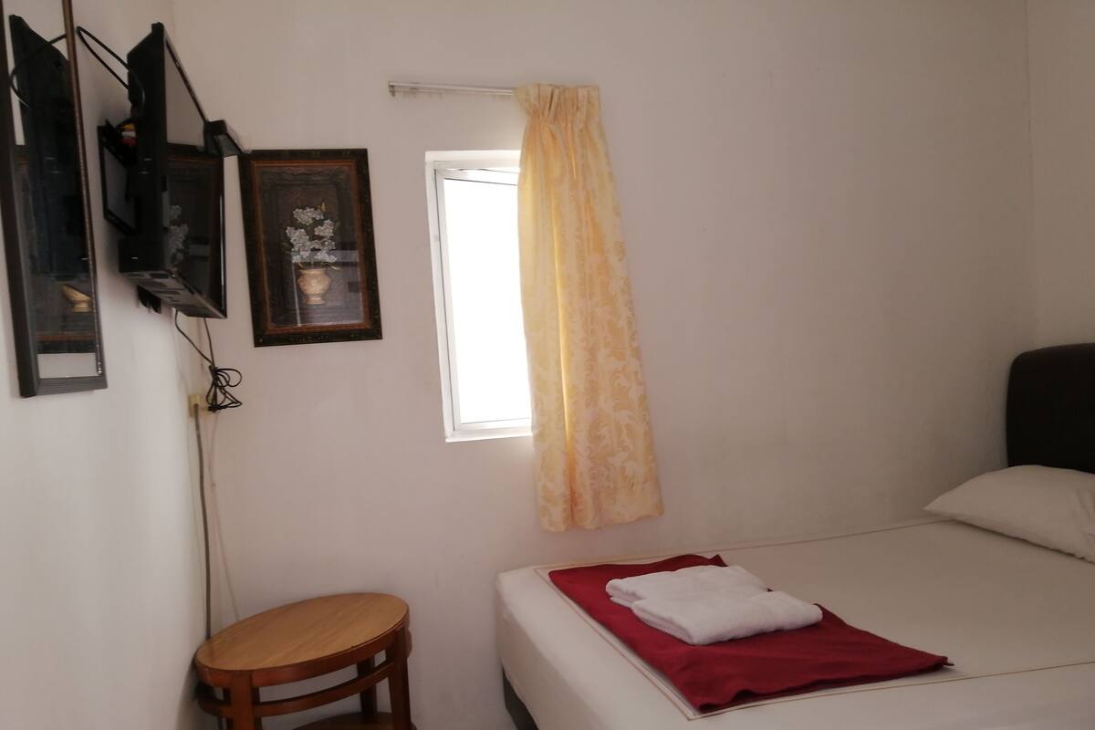 A bedroom with a bed, a small wooden stool, a window with a curtain, and a wall-mounted TV.