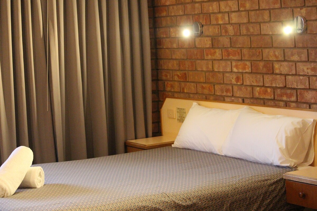 A hotel room with a brick-patterned wall, a bed with a patterned bedspread, a nightstand, and a curtain.