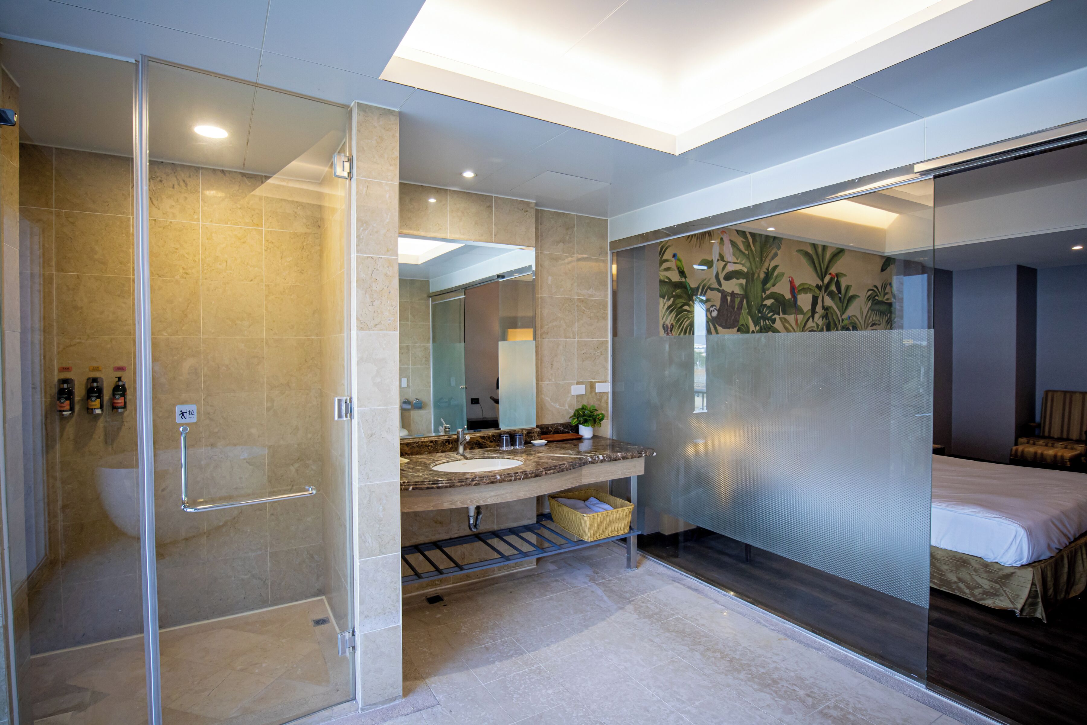 A modern bathroom with a glass shower enclosure, a double sink vanity, and a bed with a view of the room.