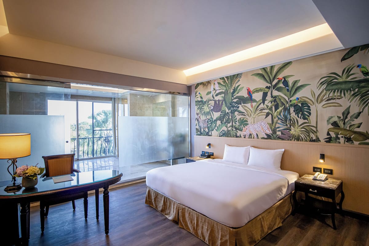 A hotel room with a large bed, a desk with a lamp, a chair, a small table, and a balcony with a glass door.