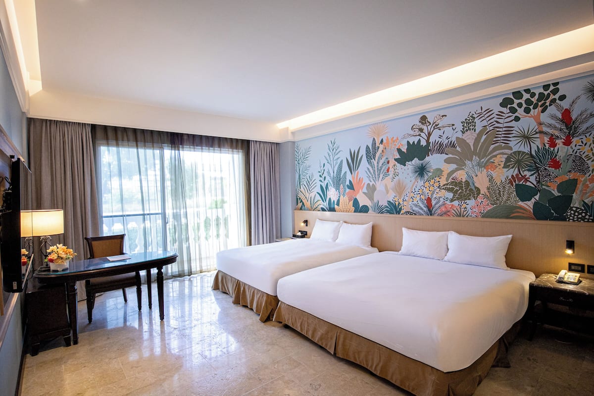 A hotel room with two beds, a desk, a chair, a lamp, and a large floral mural on the wall.