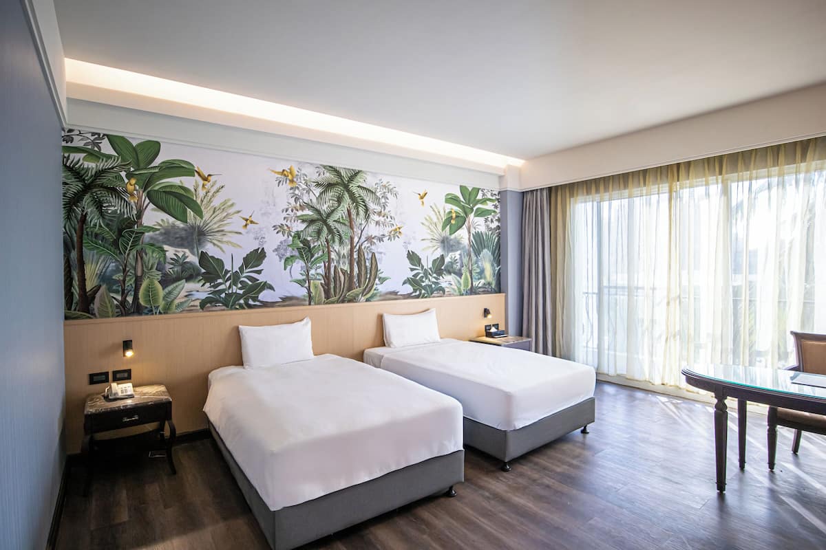 A hotel room with two beds, a mural of tropical plants on the wall, a desk with a chair, and a large window with curtains.