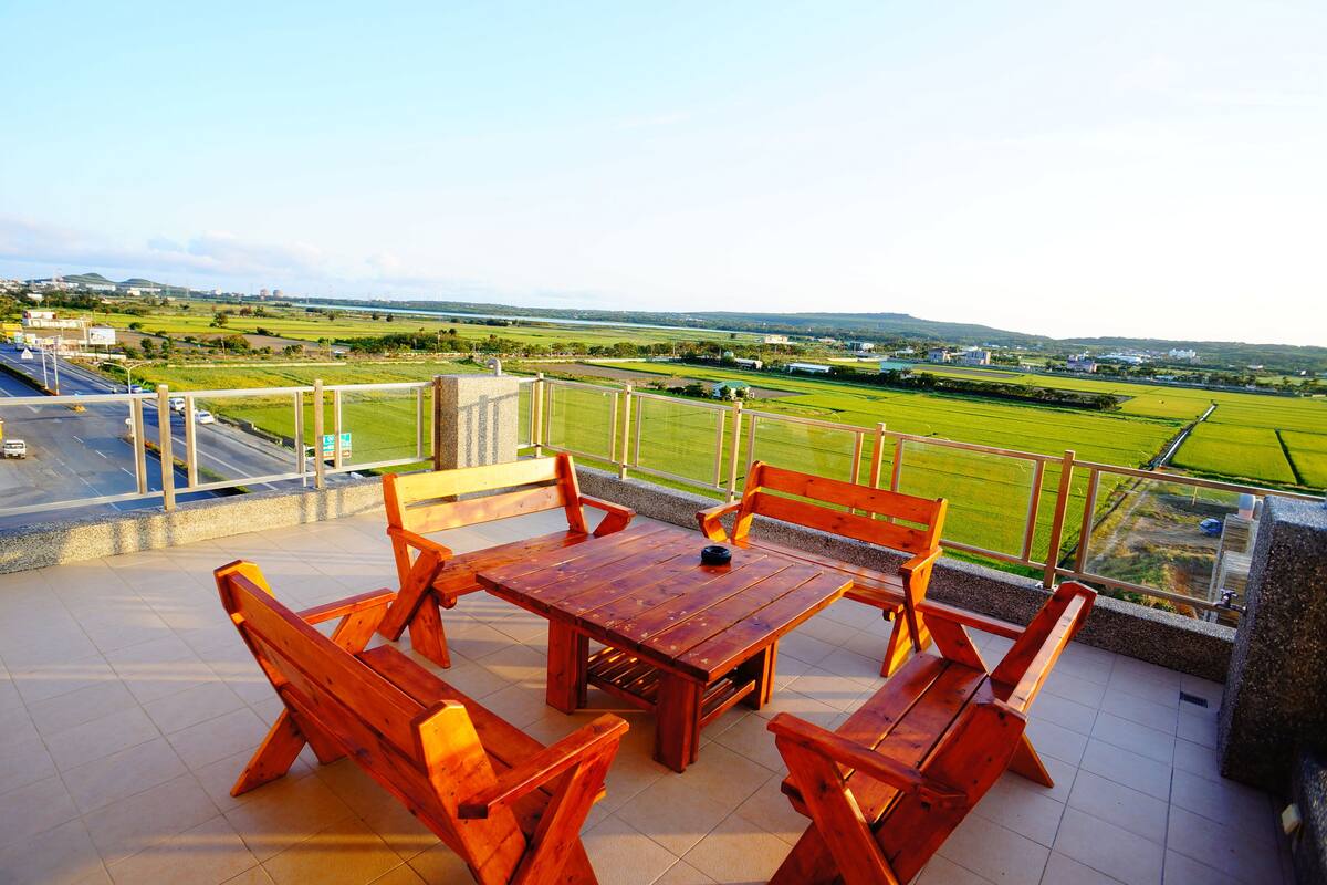 A wooden table and chairs on a balcony with a view of green fields and distant buildings.