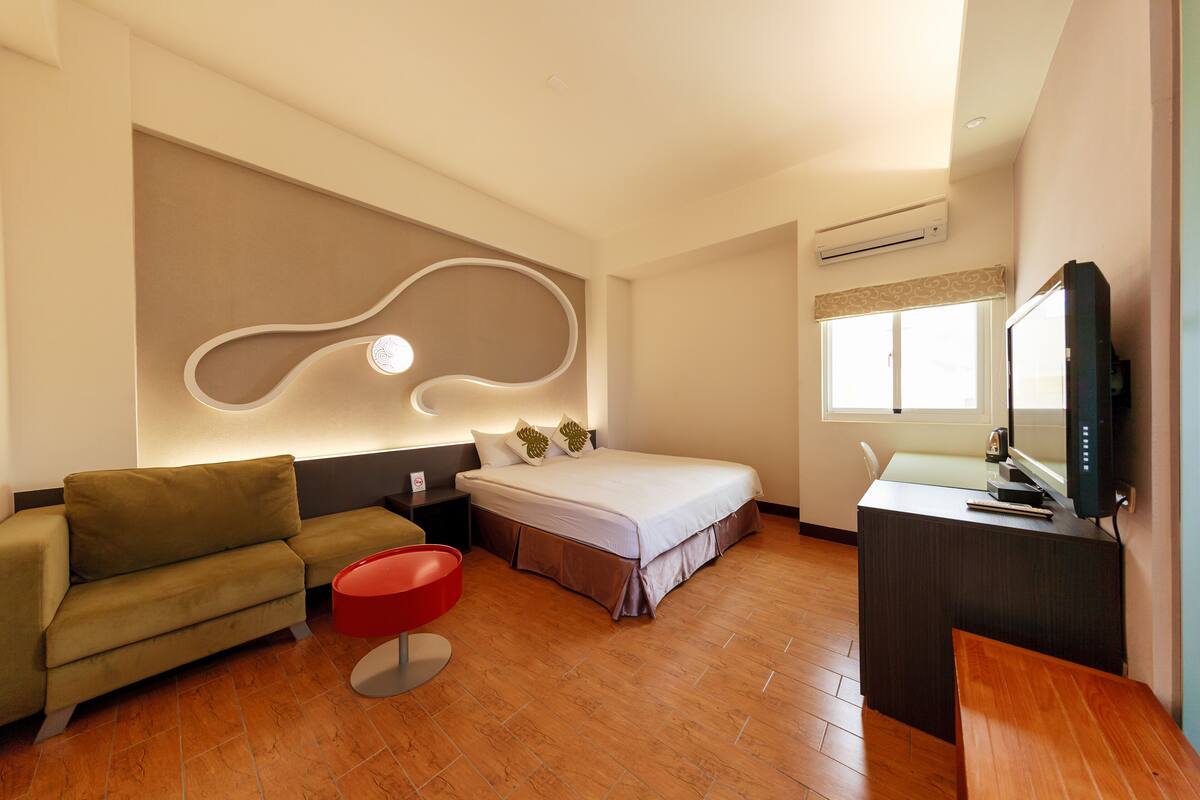 A modern hotel room with a bed, sofa, red ottoman, desk, and a television.