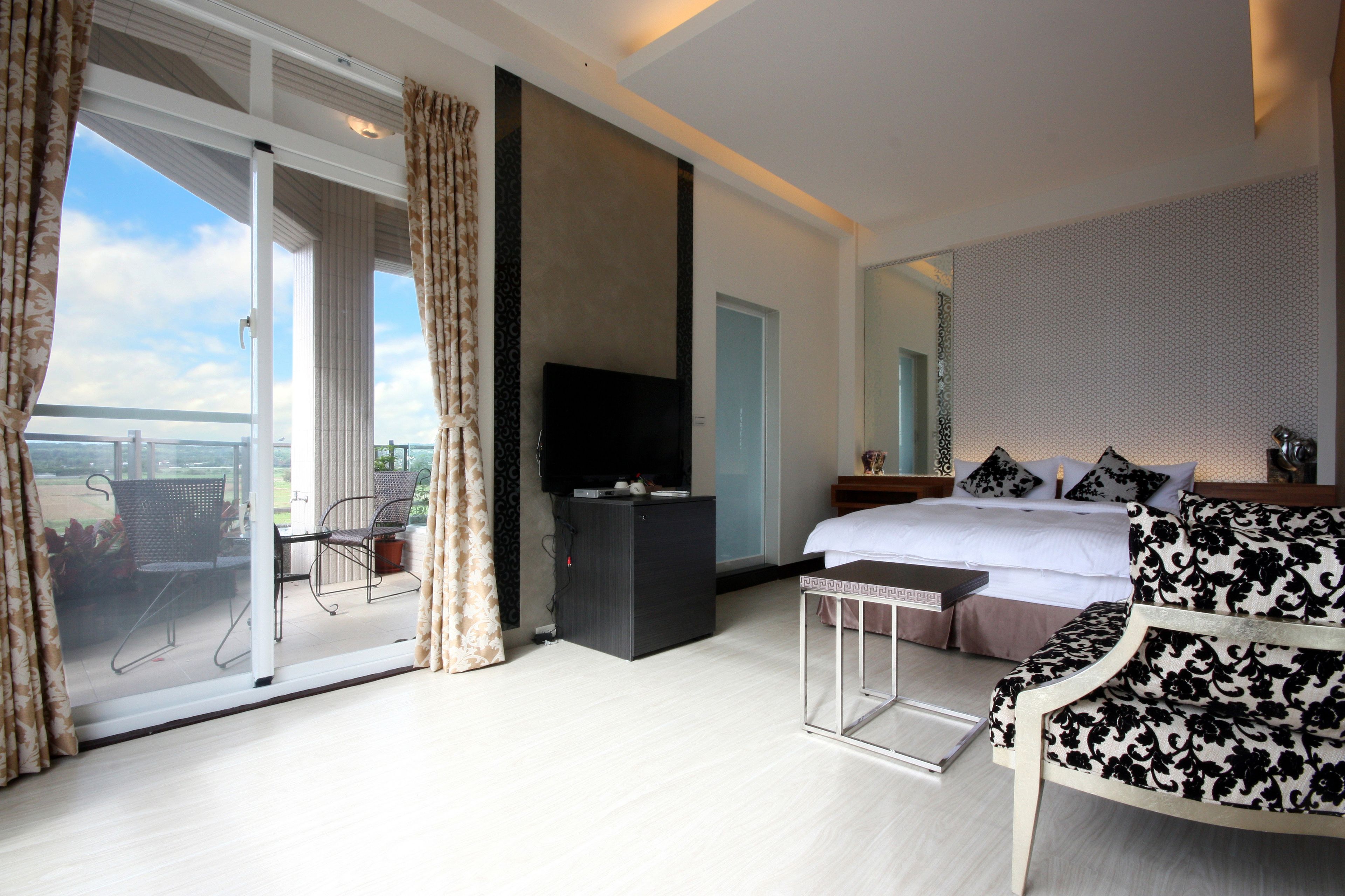 A modern hotel room with a large bed, a TV, a balcony with outdoor furniture, and a view of the city.