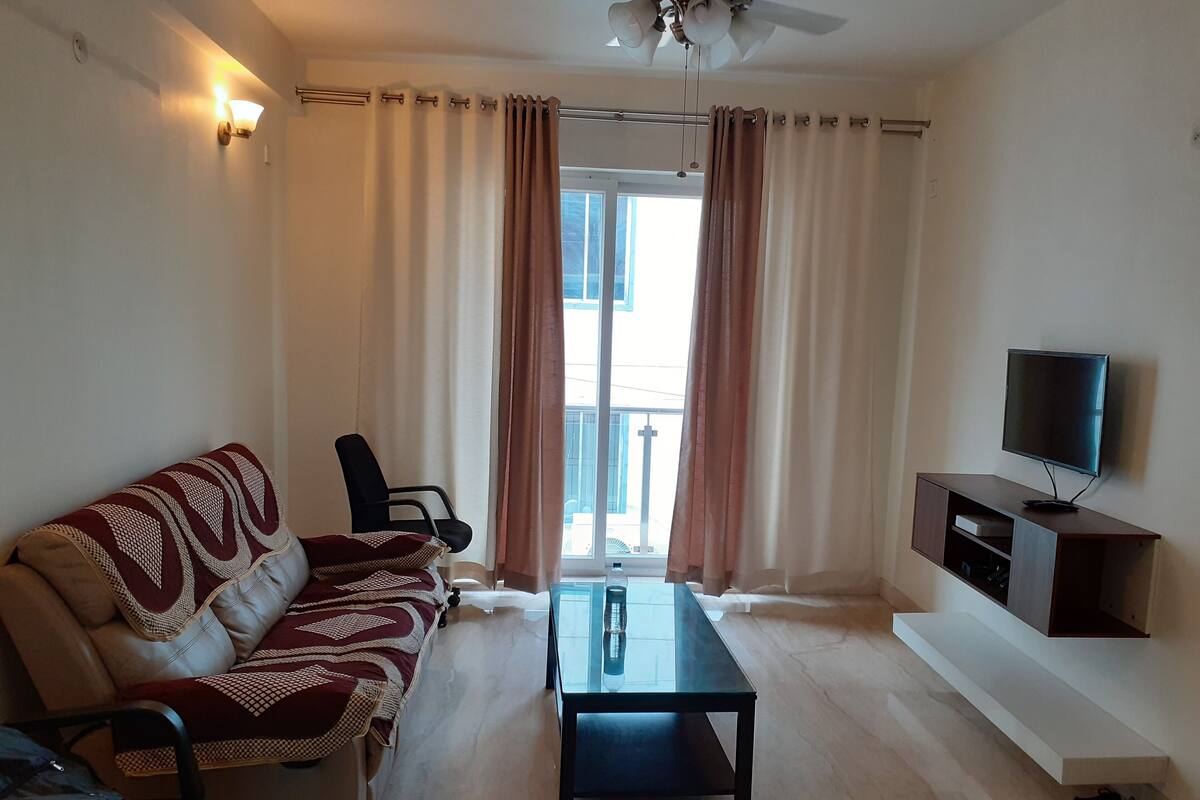 Una sala de estar con sofá, una mesa de centro, un televisor sobre un mueble bajo y un balcón con cortinas.