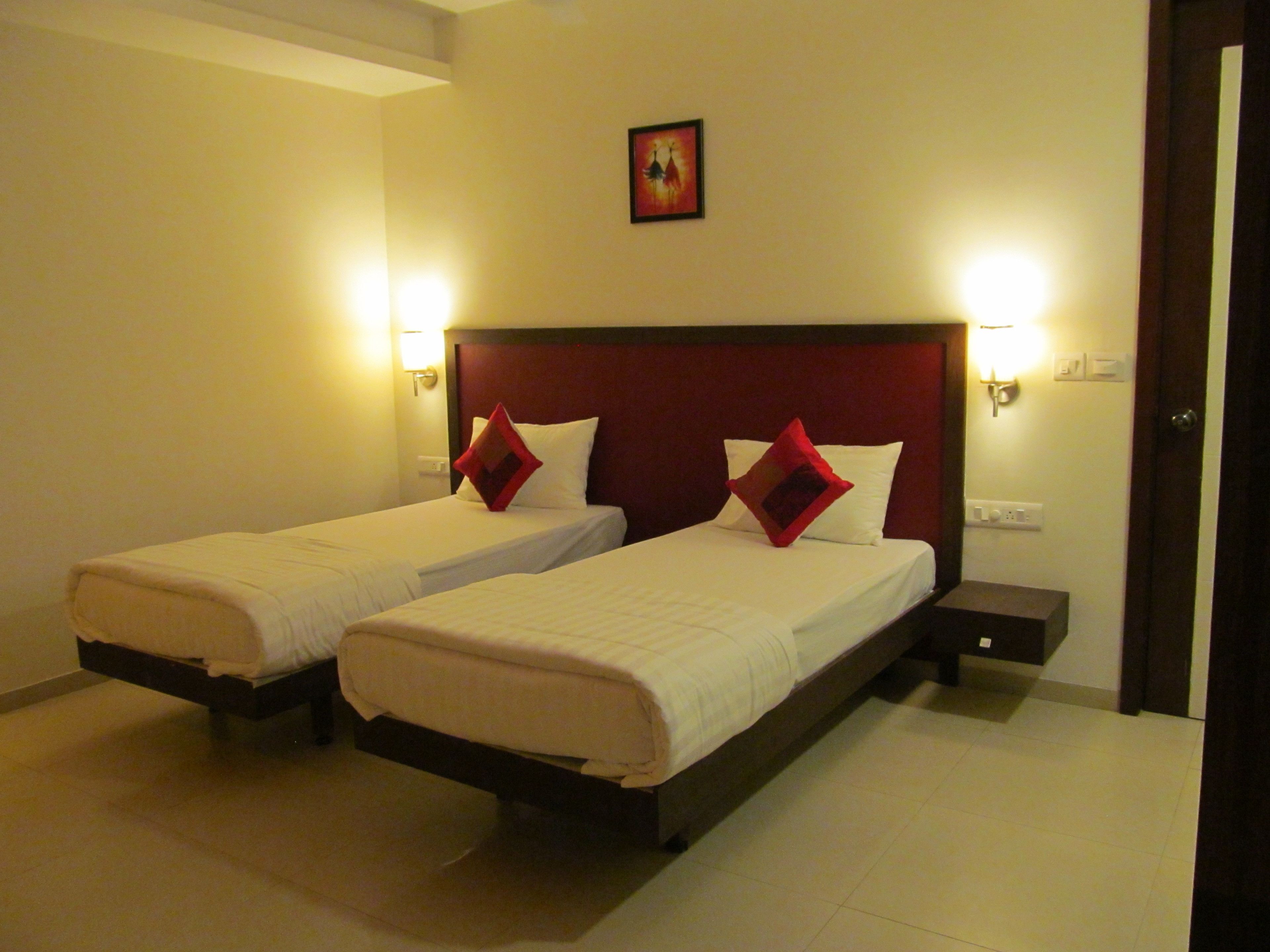 Two single beds with wooden frames and red pillows, a small bedside table, and a wall-mounted light fixture.