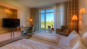 A hotel room with two beds, a desk with a television, a chair, a small table with a vase, and a view of a grassy area through a glass door.