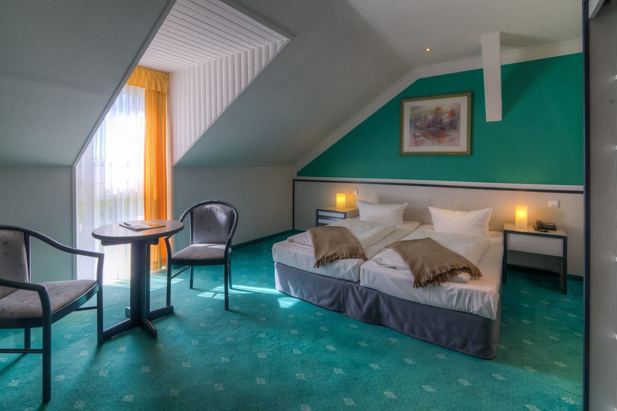 A hotel room with a bed, two bedside tables, a chair, a small table, and a window with curtains.