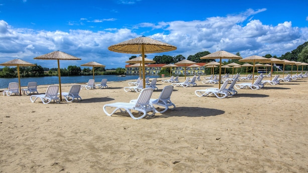A beach with white lounge chairs and thatched umbrellas, a calm body of water, and a clear sky.