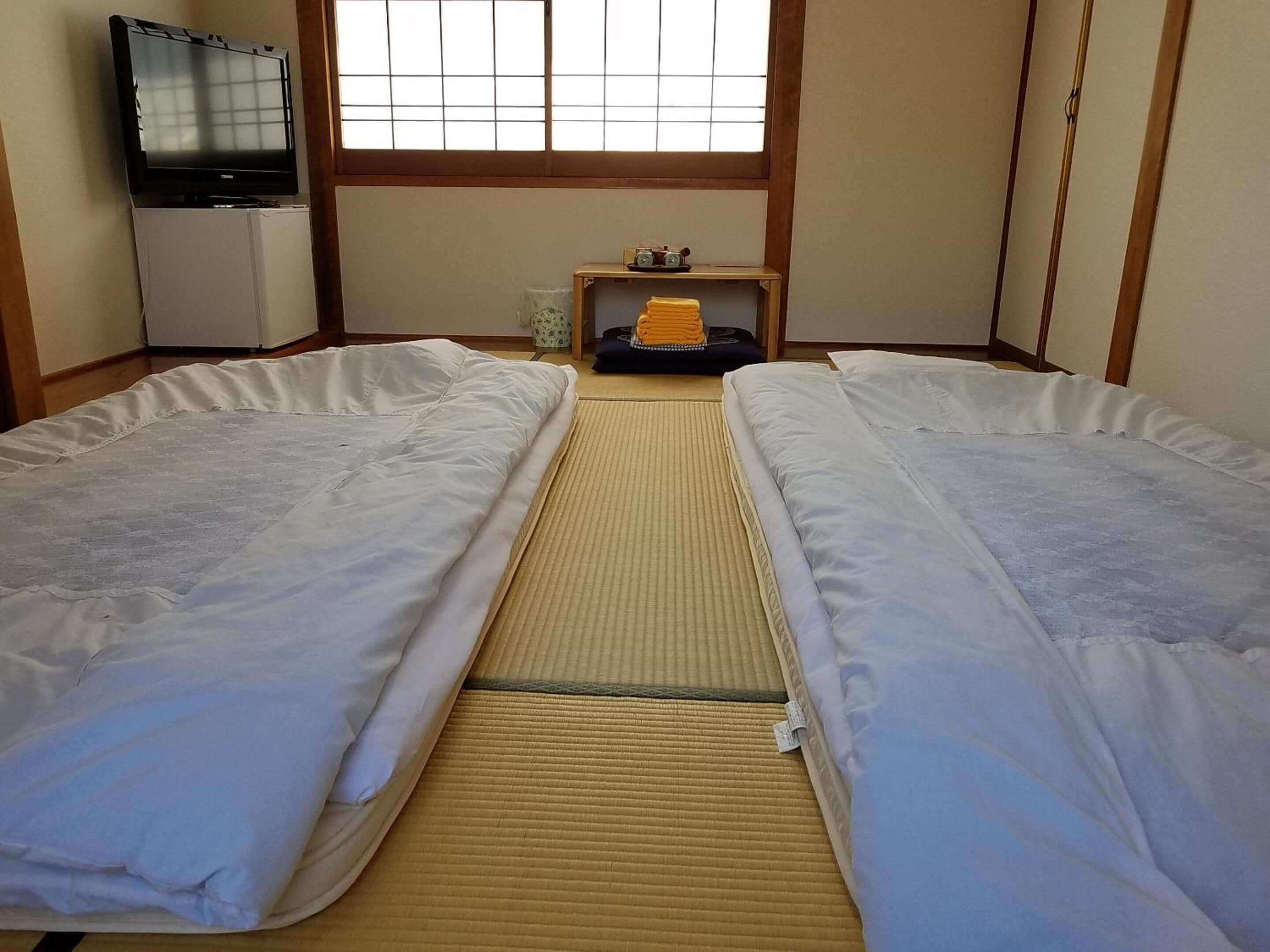 A room with two beds, a television, a small table with a heater, and a window with a grid pattern.