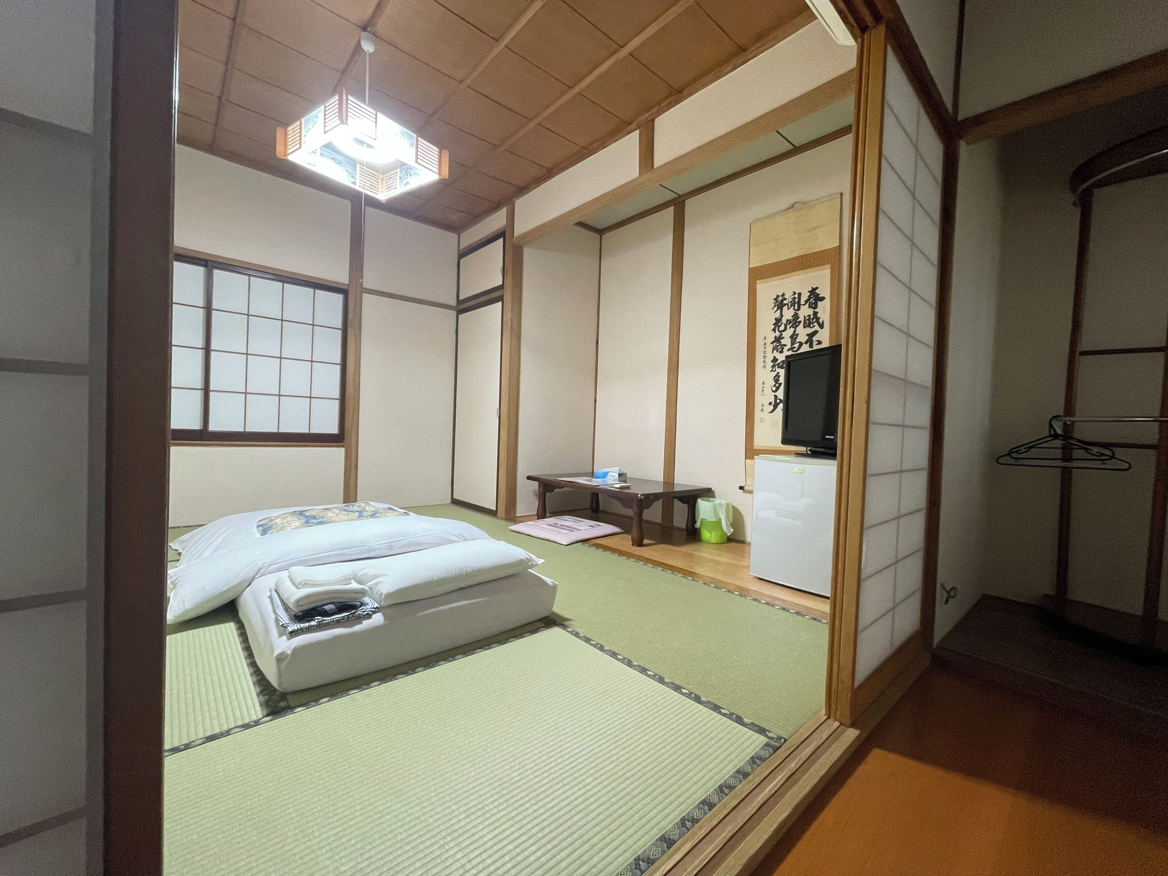 Een traditionele Japanse kamer met tatami vloeren, een bed met witte beddengoed, een televisie en een klein tafeltje met een stoel.