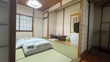 Een traditionele Japanse kamer met tatami vloeren, een bed met witte beddengoed, een televisie en een klein tafeltje met een stoel.