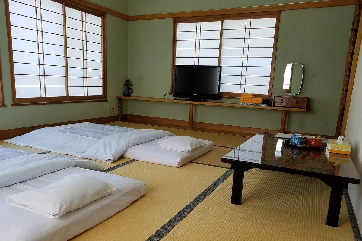 Ein traditioneller japanischer Raum mit Tatami-Matten, zwei Betten, einem Fernseher, einem kleinen Tisch mit Teeservice und einem Spiegel.
