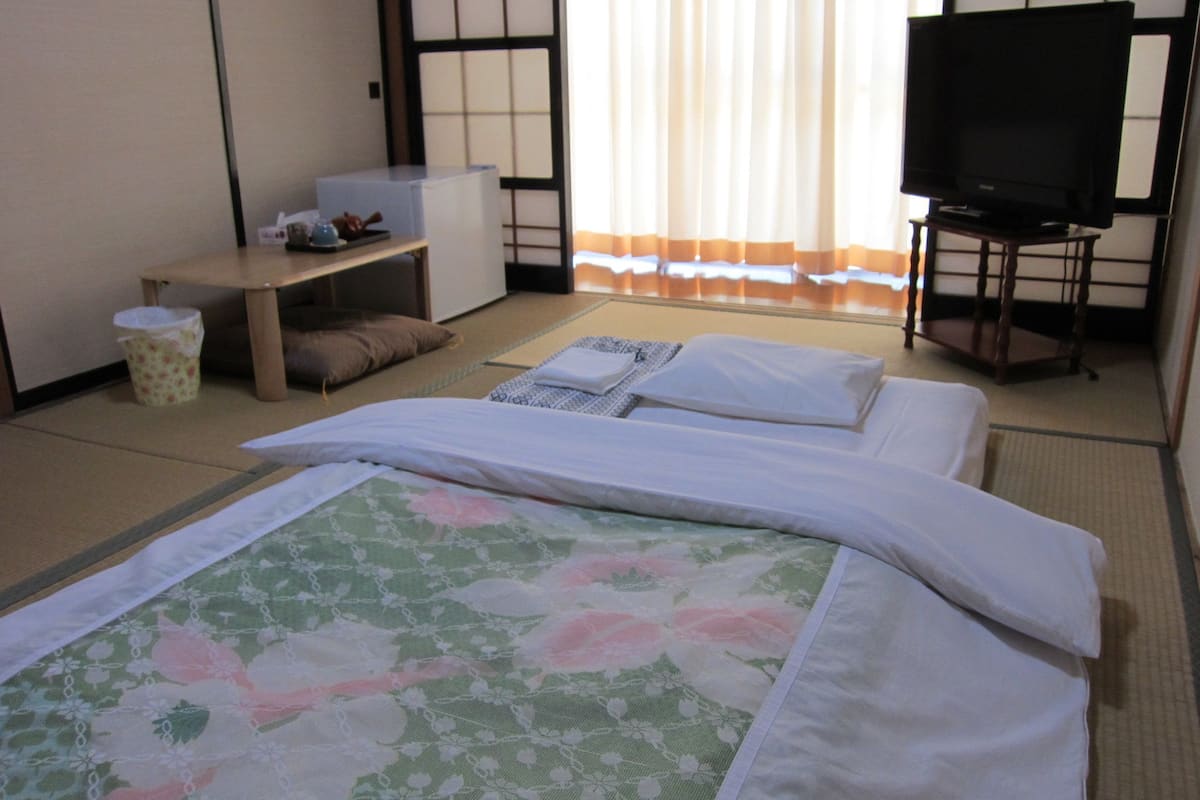 Ein Hotelzimmer mit Tatami-Boden, einem Bett mit floralem Bettbezug, einem Fernseher auf einem Schrank und einem kleinen Tisch mit einem Papierkorb.