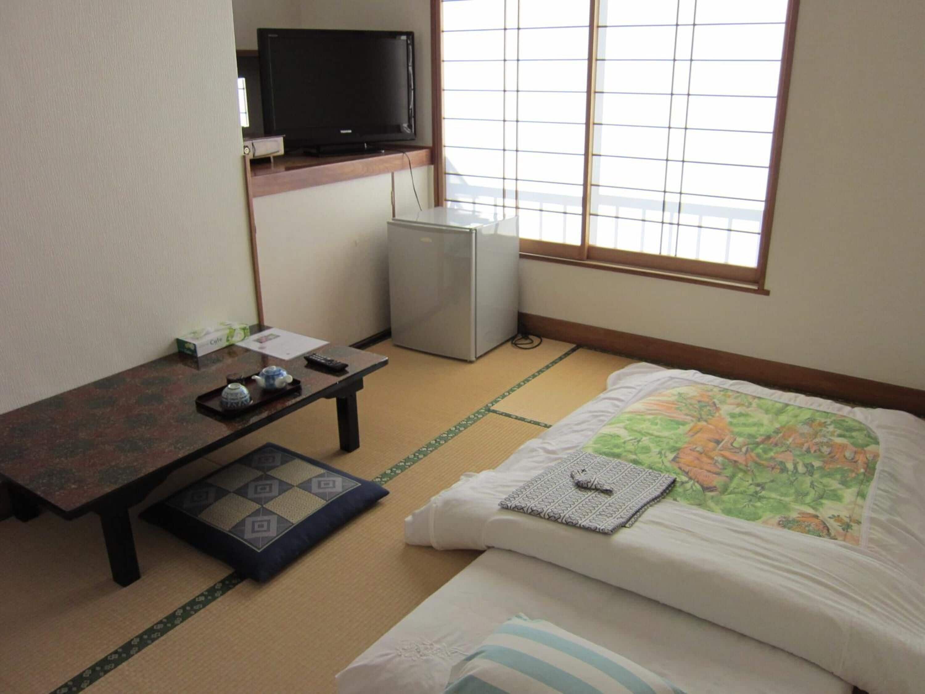 A hotel room with tatami flooring, a bed with a floral bedspread, a small table with a teapot and remote control, a television on a shelf, and a refrigerator.