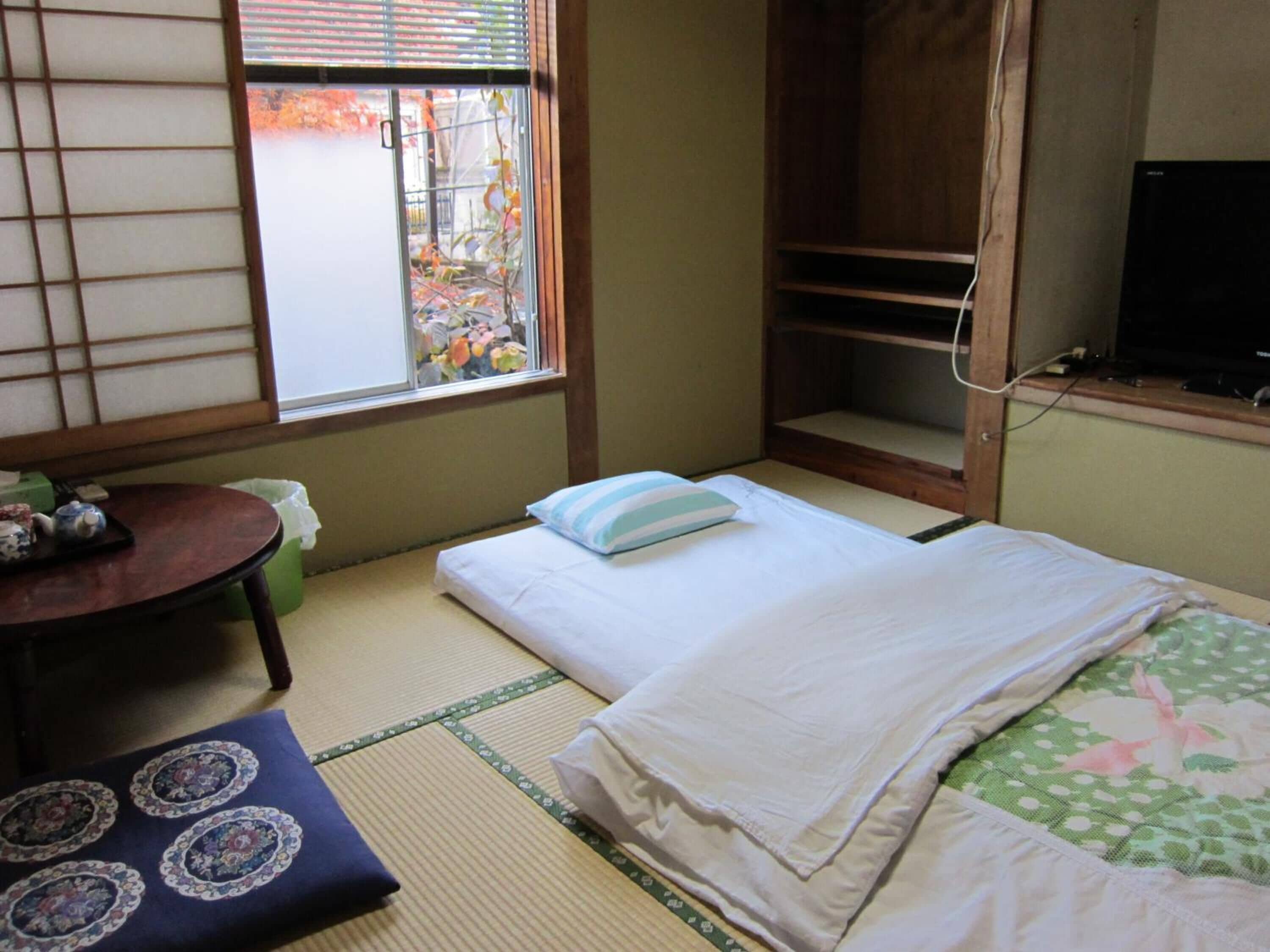 Una habitación tradicional japonesa con suelo de tatami, una cama futón, una mesita con tetera y un televisor sobre una estantería de madera.