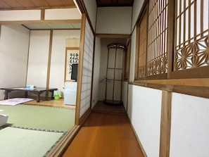 Une pièce de style japonais traditionnel, avec un sol recouvert de tatamis, une basse table en bois, un réfrigérateur blanc et une porte coulissante ornée d’un motif en treillis décoratif.