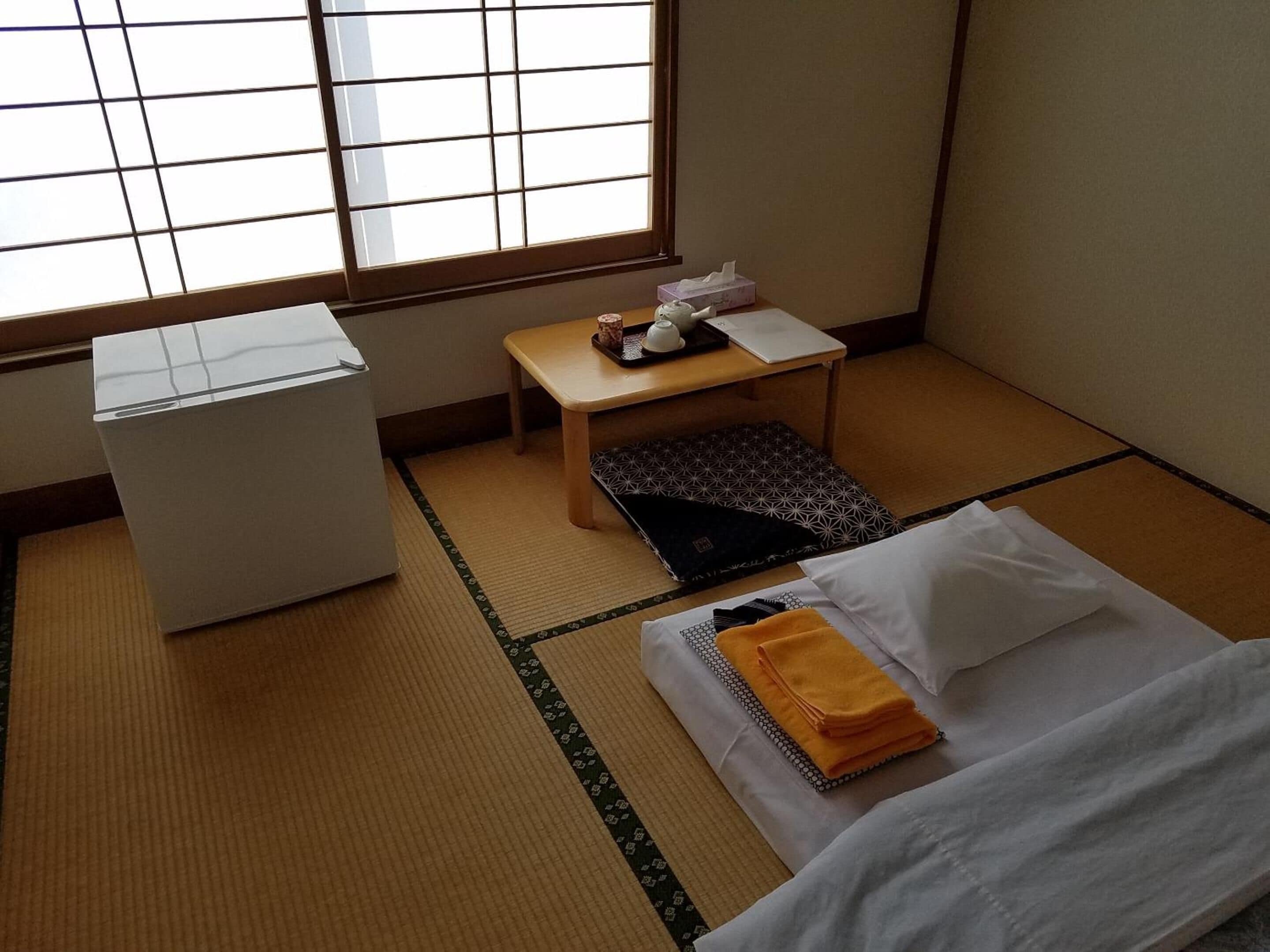 A room with tatami flooring, a bed with a white and orange blanket, a small wooden table with a tissue box and a cup, and a white air conditioning unit.