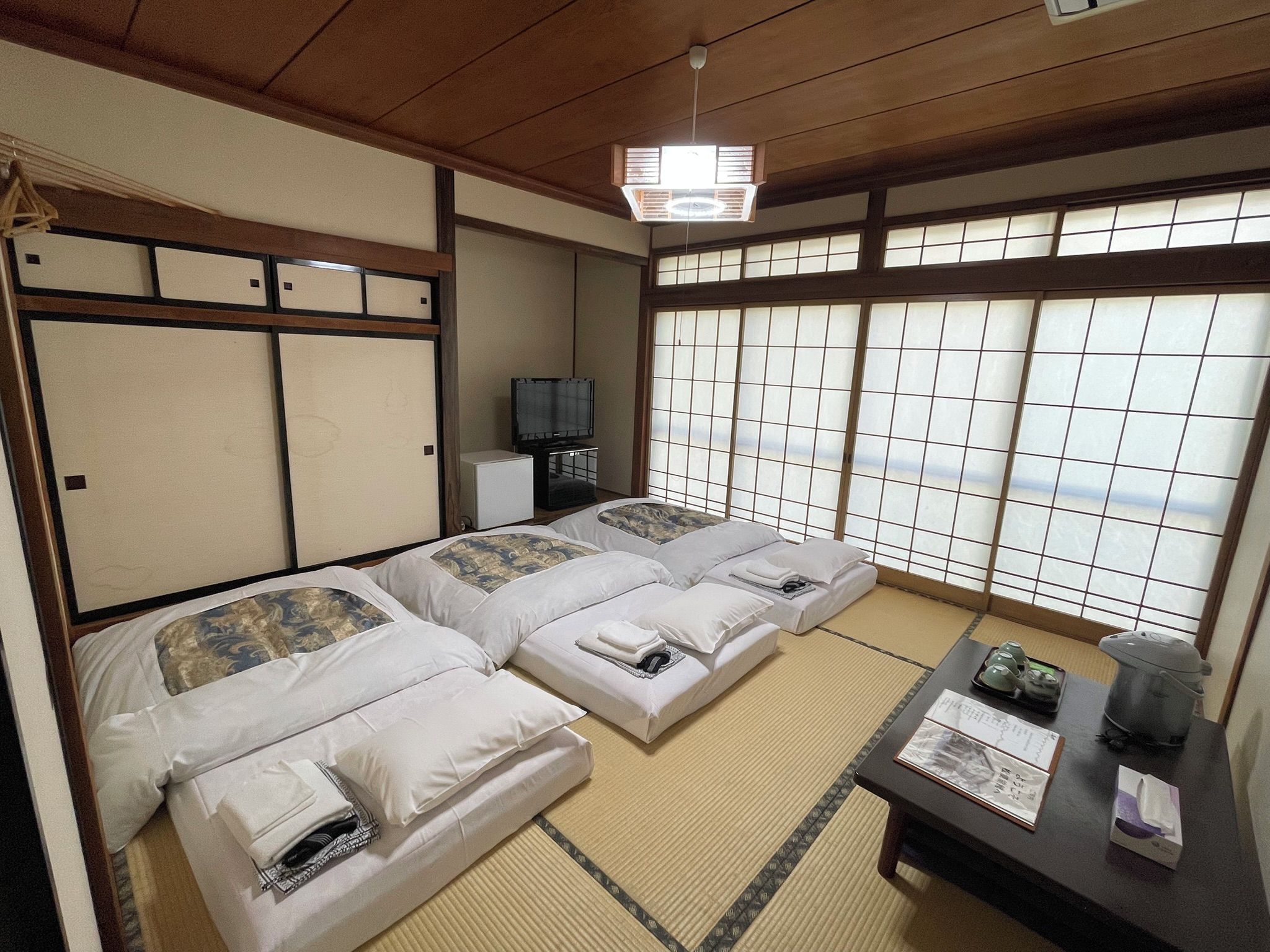 A traditional Japanese-style room with tatami flooring, two beds with pillows, a television, and a view through shoji screens.
