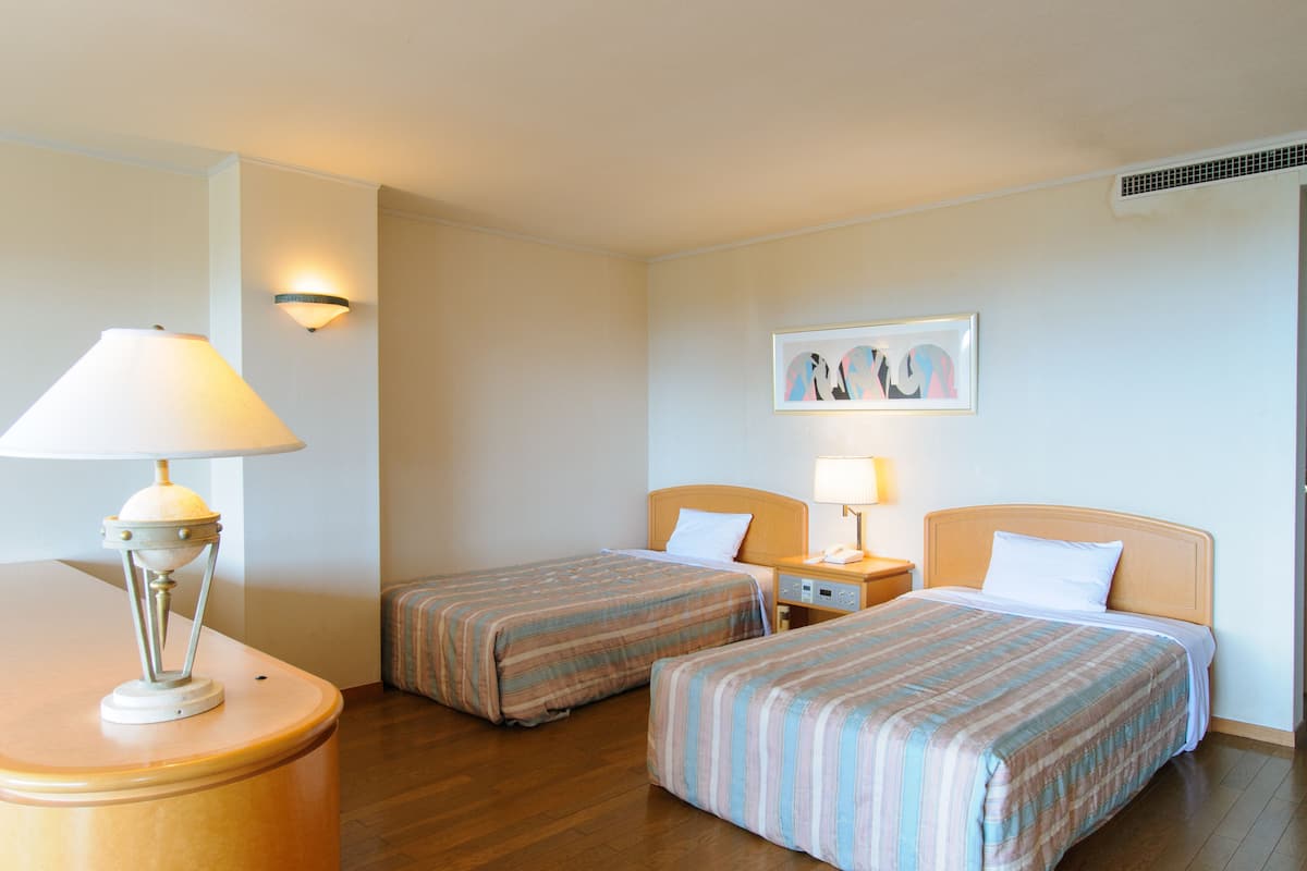 A hotel room with two single beds, a nightstand with a lamp, a wall-mounted artwork, and a wooden floor.