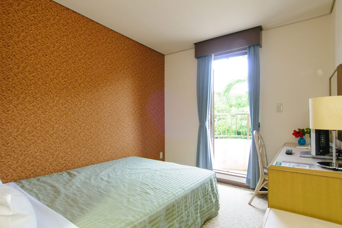 A hotel room with a bed, a desk with a lamp, a chair, and a view of the outdoors through a window with curtains.