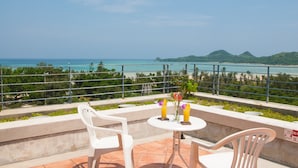 Un balcon avec deux chaises en plastique blanches, une petite table ronde et deux boissons. Le balcon donne sur une plage et l’océan.
