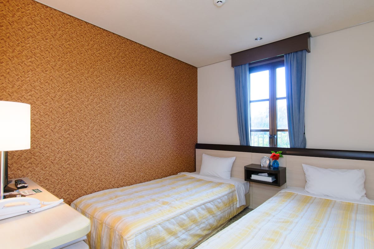 A hotel room with two beds, a nightstand, a lamp, and a window with curtains.