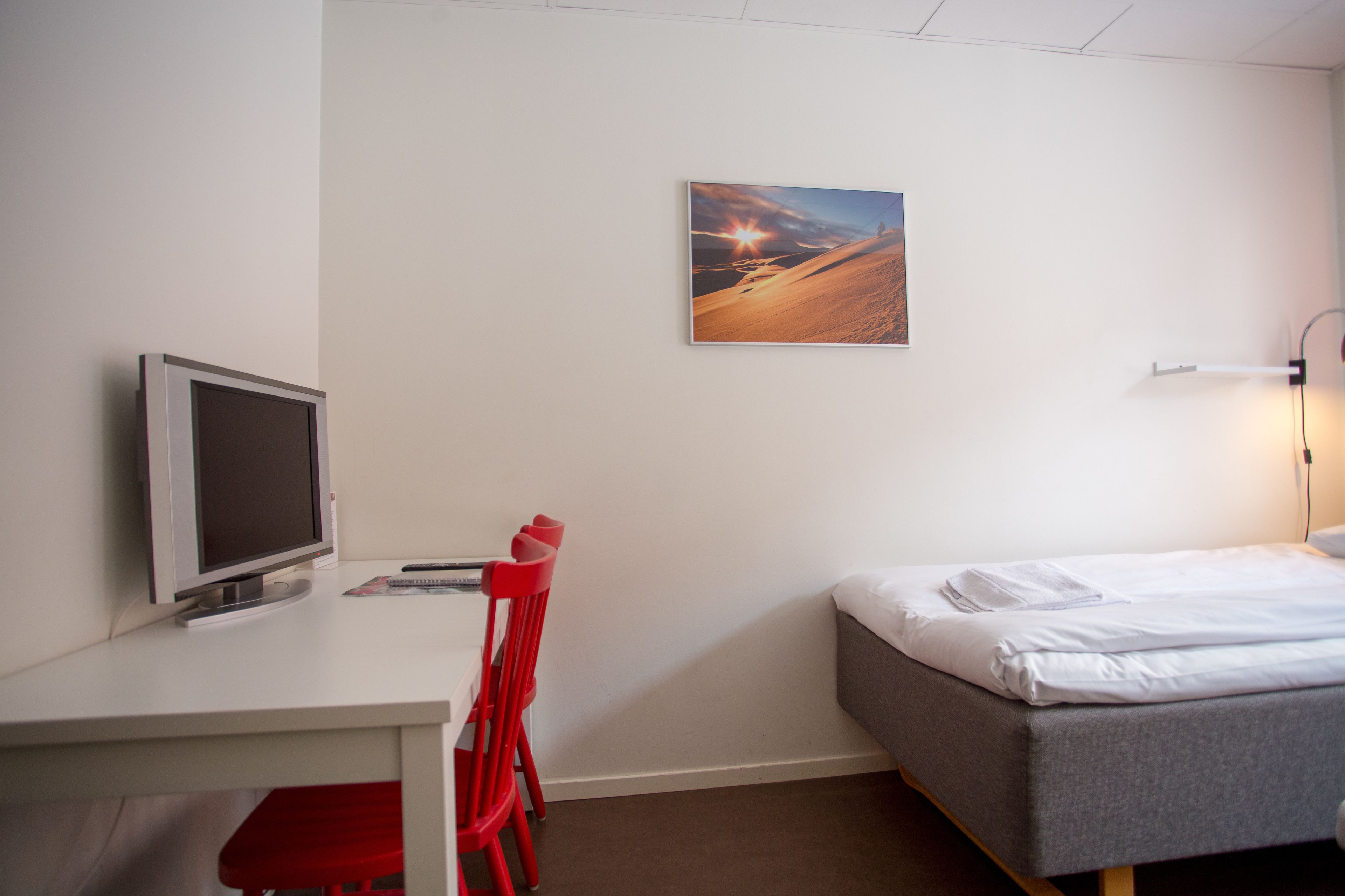 A room with a bed, a desk with a computer monitor, a red chair, and a wall-mounted picture.