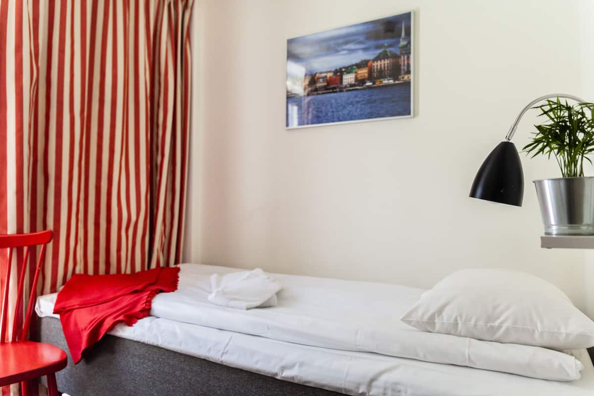 Una camera d'albergo con un letto, una poltrona rossa, una coperta rossa, un cuscino bianco, una lampada nera, una pianta in vaso di metallo e un dipinto con una veduta cittadina sulla parete.