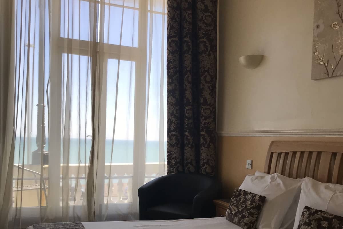 A hotel room with a large bed, a chair, and a view of the sea through sheer curtains.