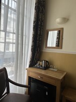 A hotel room with a wooden desk, a refrigerator, a coffee maker, and a mirror on the wall.