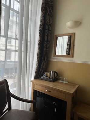 A hotel room with a wooden desk, a refrigerator, a coffee maker, and a mirror on the wall.