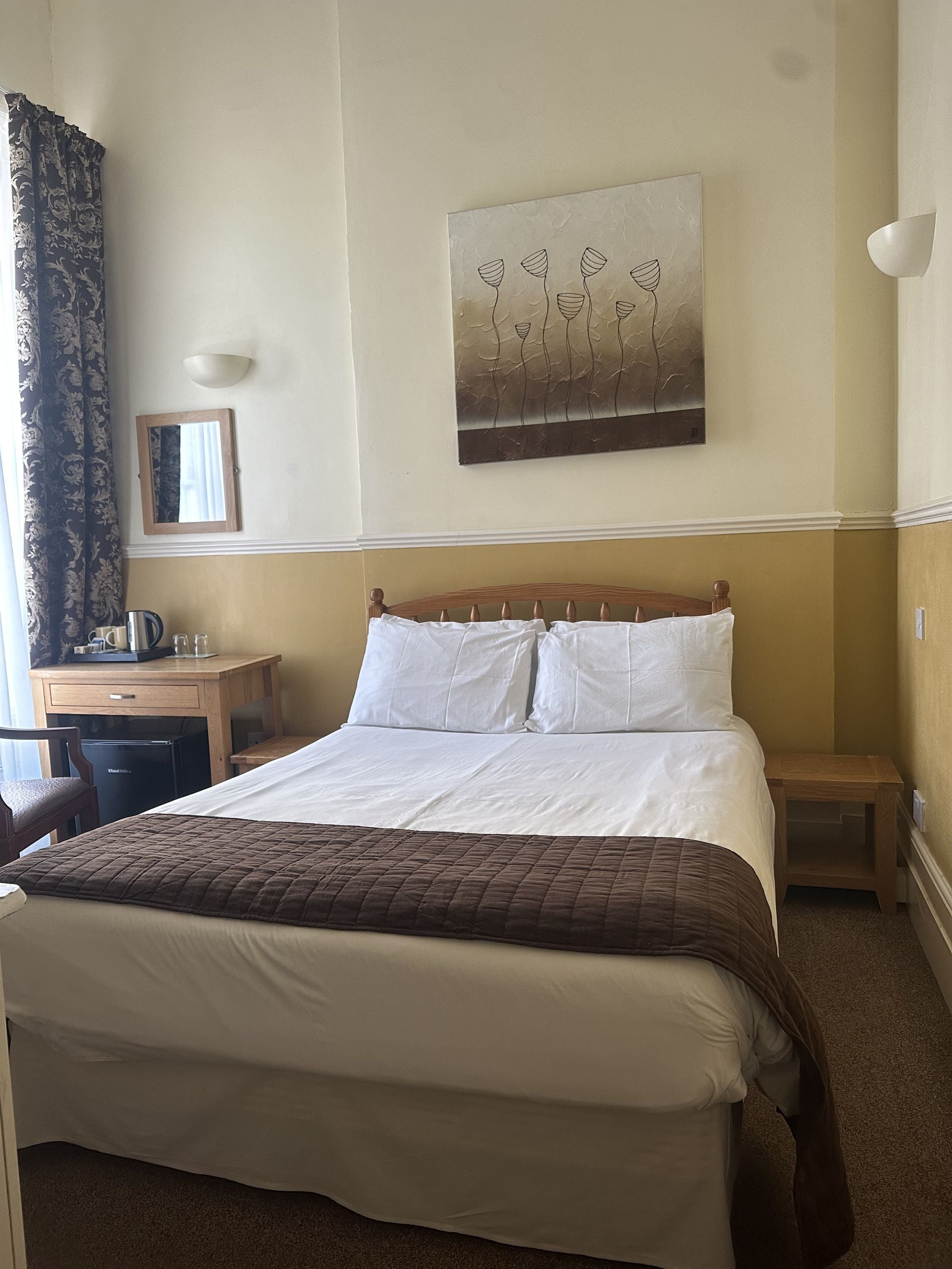 A hotel room with a bed, a desk, a chair, a small table, a lamp, and a painting on the wall.