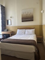A hotel room with a bed, a desk, a chair, a small table, a lamp, and a painting on the wall.