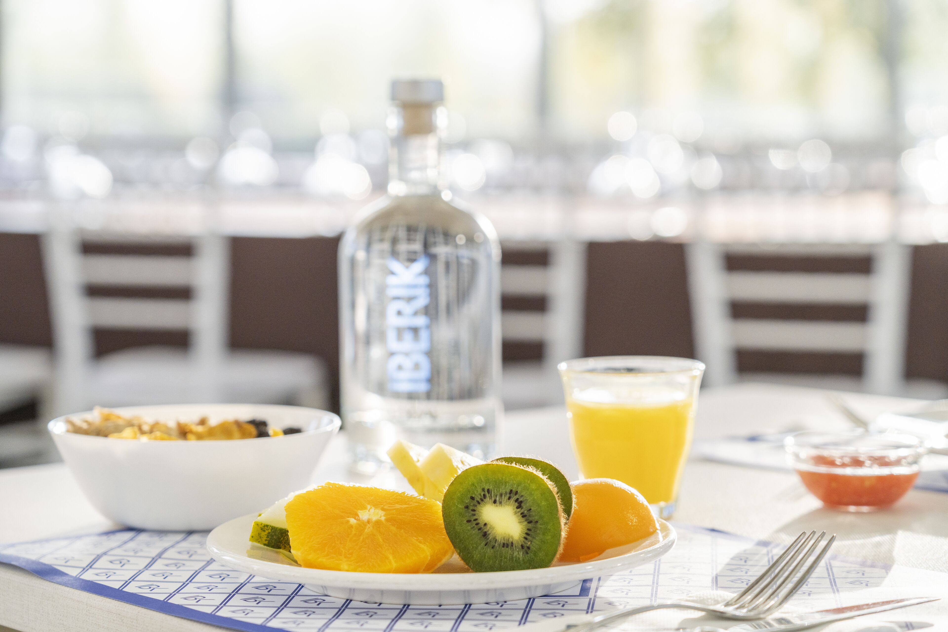 A breakfast setting with a plate of fruit, a glass of orange juice, a bottle of water, and a bowl of cereal.