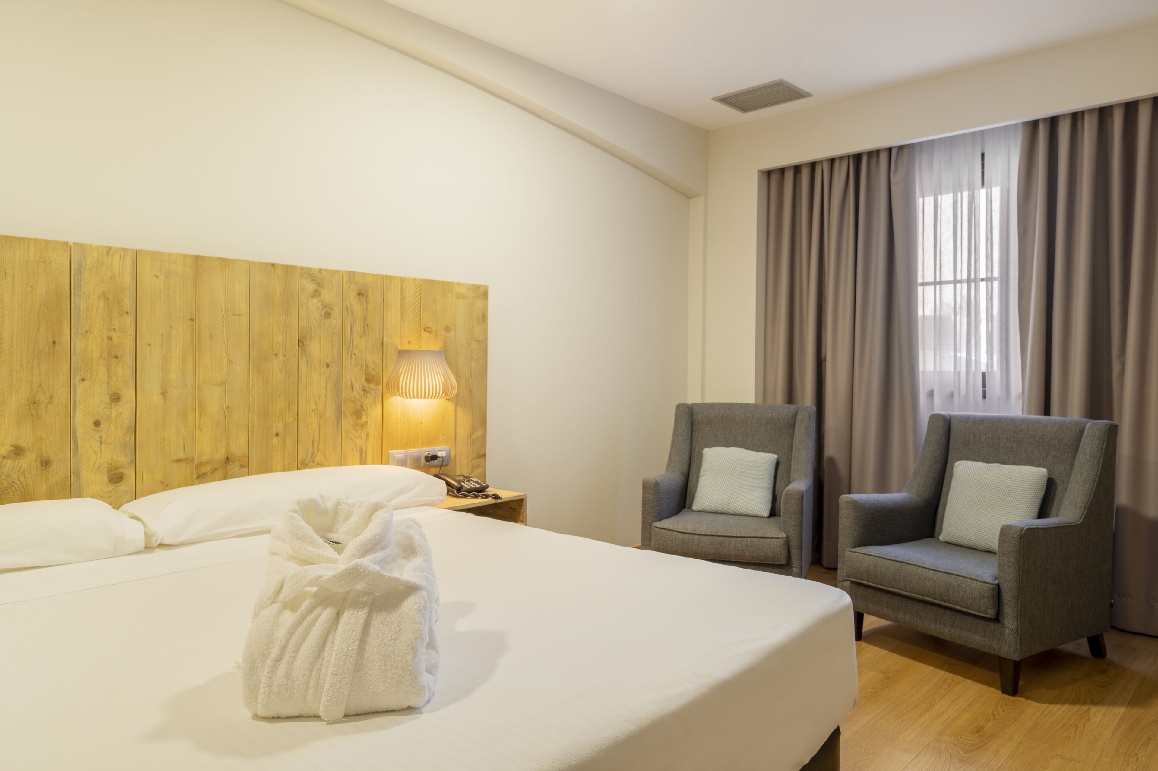 A hotel room with a large bed, two armchairs, a wooden headboard, a nightstand with a lamp, and a window with curtains.