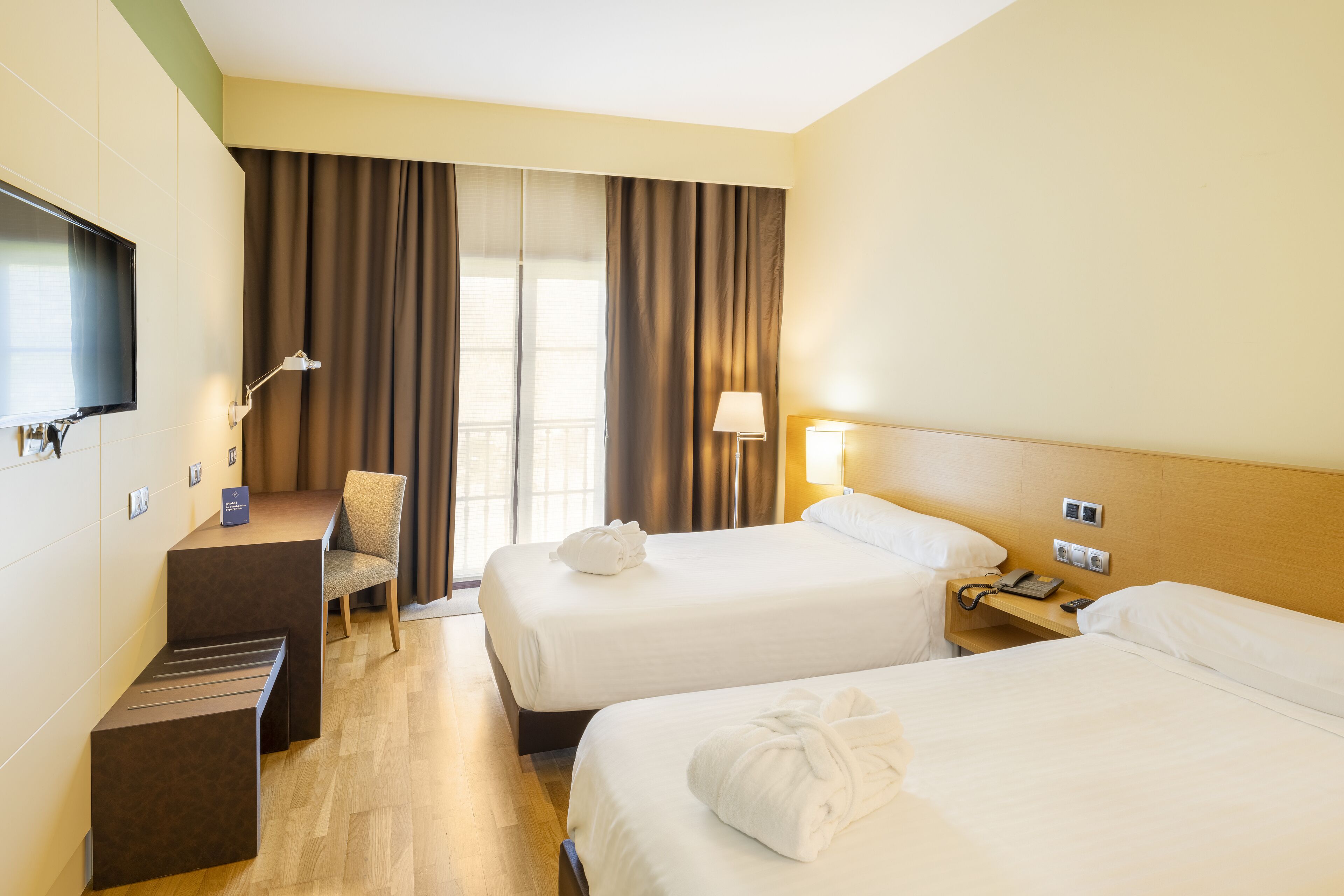 Une chambre d’hôtel avec deux lits, un bureau, une chaise, une télévision et une fenêtre avec des rideaux.
