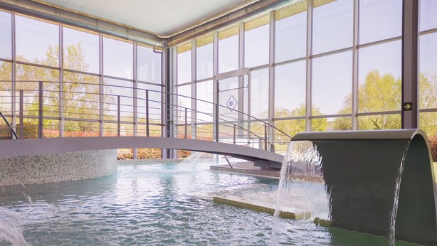 Una piscina interior con cascada, ventanas amplias y vistas a árboles fuera.