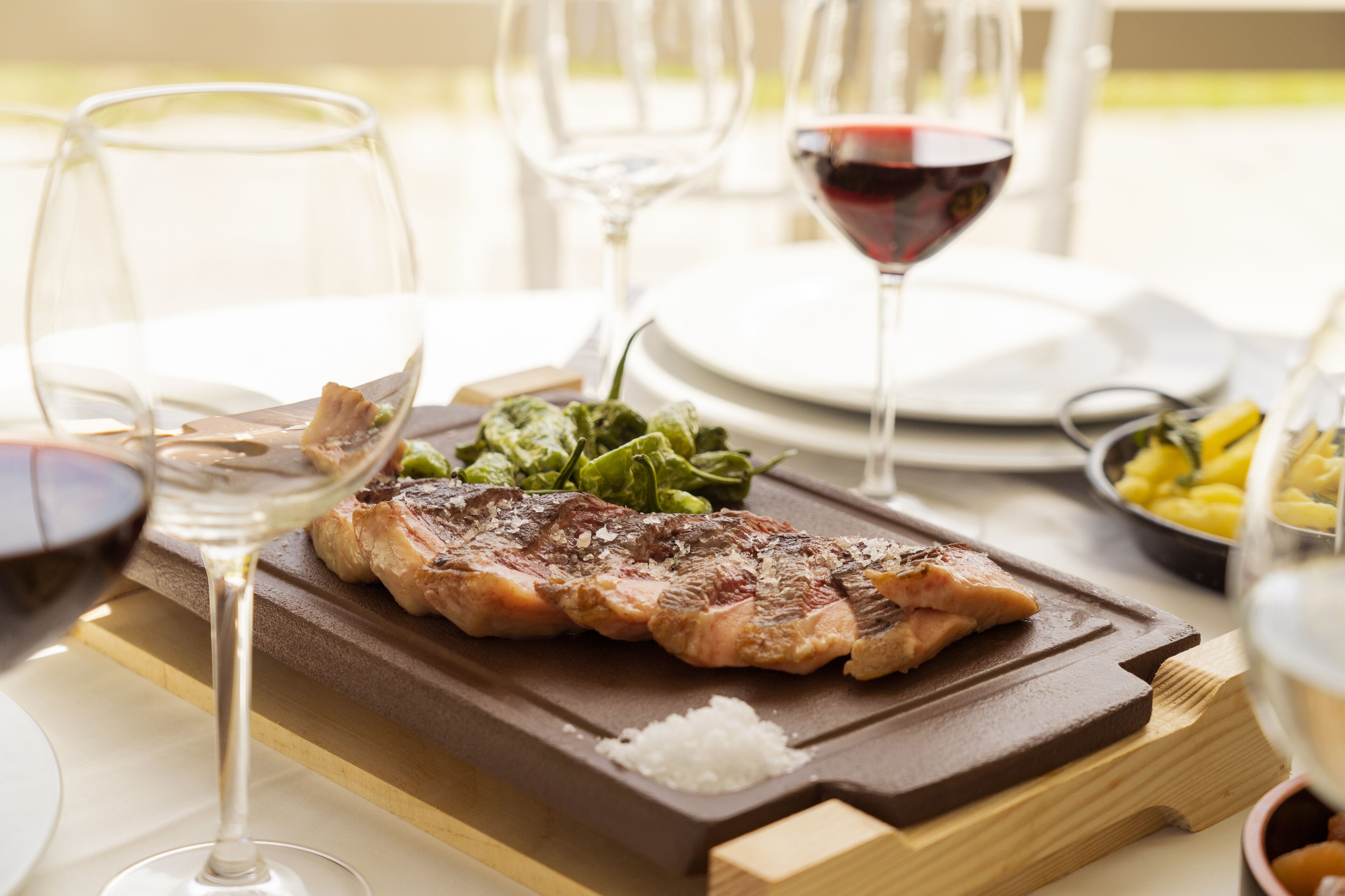 A steak on a wooden board with a side of greens, a glass of red wine, and a bowl of fruit on a table.