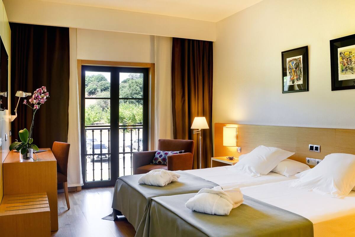 Ein Hotelzimmer mit zwei Betten, einem Schreibtisch mit Stuhl, einer Schreibtischlampe, einer Zimmerpflanze und Blick ins Freie durch ein großes Fenster.