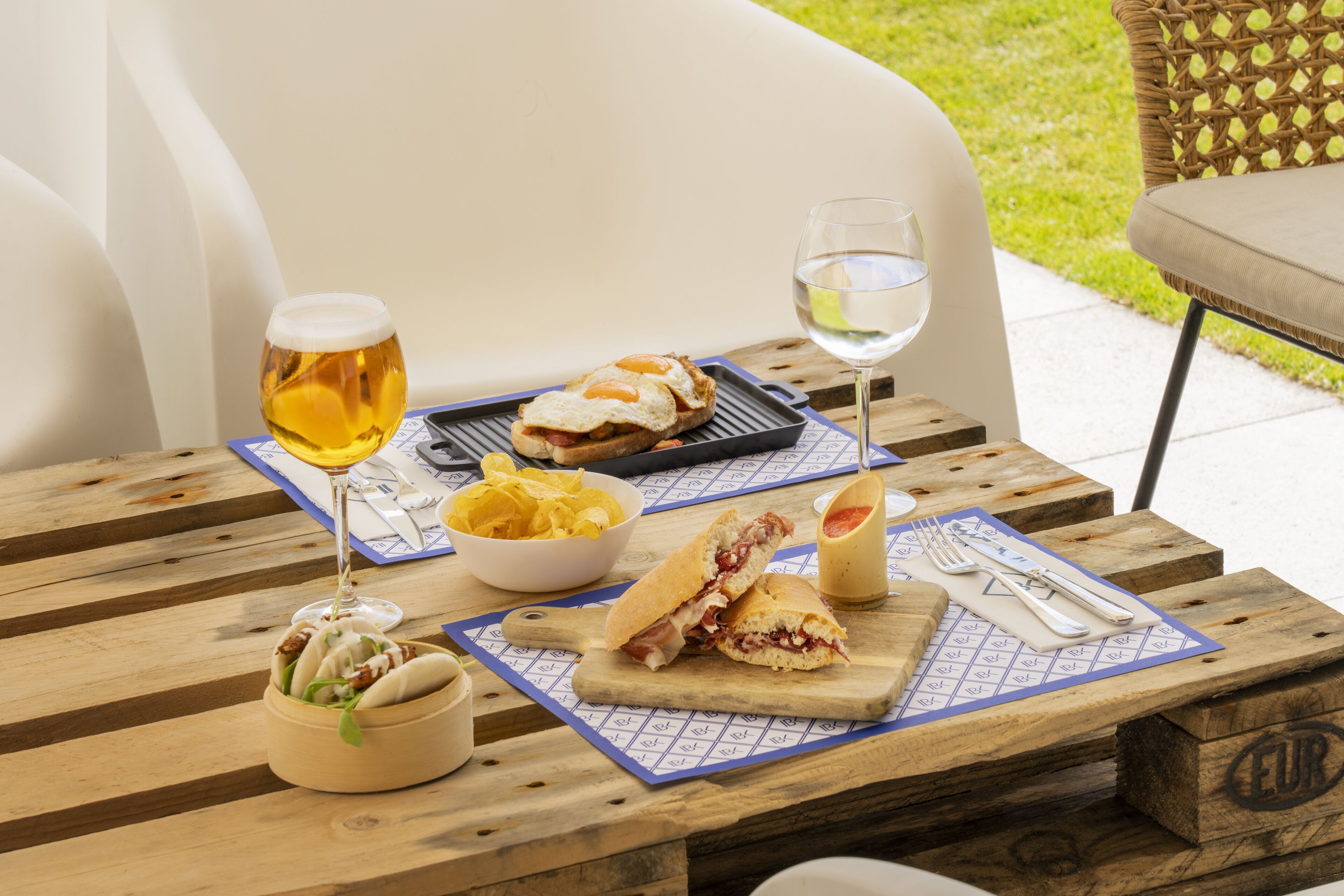 Ett träbord med en smörgås, en skål med chips, ett glas öl och ett glas vatten.